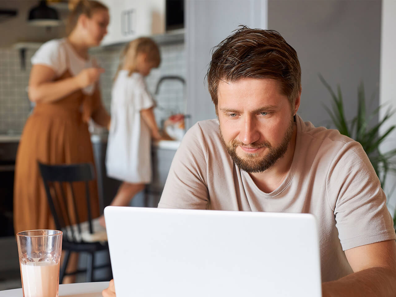 Ein junger Mann sitzt am Laptop. Im Hintergrund sind seine Frau und sein Kind zu sehen.